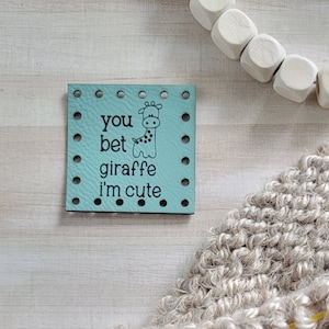 May include: A close-up of a beige knitted fabric with a wooden knitting needle and a pink flower embellishment. The fabric is being worked on with a yellow rubber band. A blue card with the text "you bet giraffe i'm cute" is in the background.