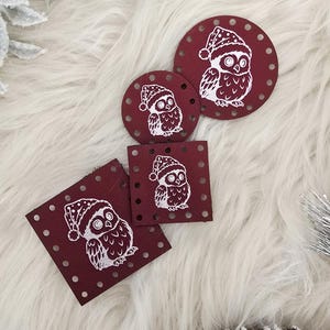 May include: A collection of burgundy-colored, circular and square ornaments with a white owl design wearing a winter hat. The ornaments are arranged on a white, fluffy surface, with decorative winter foliage and a pinecone in the background.