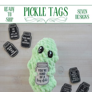 May include: Seven different pickle-themed gift tags with various sayings. The tags are printed on white cardstock and shaped like mason jars. The tags are displayed on a wooden surface with a green crocheted pickle plush toy.