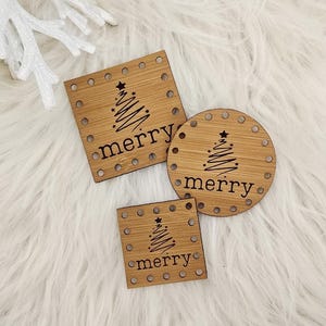 May include: Three wooden Christmas ornaments in square and round shapes, with the word "merry" and a Christmas tree design etched into the wood. The ornaments have small holes around the edges. The background is a fluffy white surface.