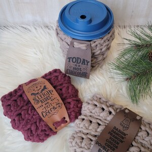 May include: Three crocheted mug cozies with different designs and text. The first cozy is maroon with the text "Old Fashioned Sleigh Rides and Hot Cocoa". The second cozy is beige with the text "Today is a Hot Chocolate". The third cozy is beige with the text "Dashin' Dancin' Prancin' Vixen Cocoa Cider Double Shot Blitzen Redbull".
