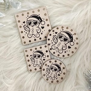 May include: Four wooden Christmas ornaments with a gingerbread man design. The ornaments are in square and circular shapes, with a black and white illustration of a gingerbread man wearing sunglasses and a Santa hat. The ornaments are decorated with small black polka dots.