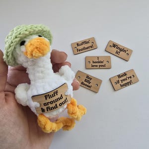 May include: A handmade white goose plushie with a green hat and yellow feet and beak. The goose has a wooden sign that says "Fluff around & find out!". There are five wooden tags with phrases like "Rufflin' feathers!" and "Honk if you're happy!"