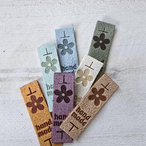 May include: A collection of rectangular fabric labels in various colors including mustard, purple, light blue, sage green, and cream. Each label features a printed flower icon, a horizontal line, and the text "hand made" in a dark brown font.