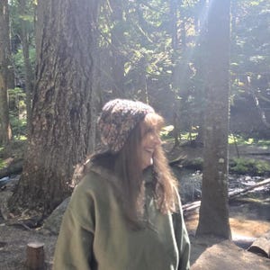 May include: A multi-colored knit beanie with a textured, speckled appearance worn with an olive green hooded sweatshirt. The beanie features a blend of earthy tones including brown, tan, and cream, providing a snug fit around the head.