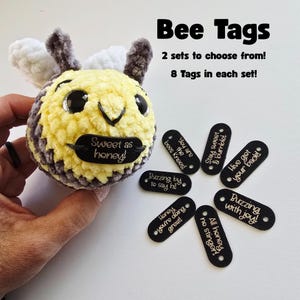 May include: A crocheted bee with a yellow and gray body, black eyes, and a black tag that says "Sweet as honey!". The image also shows black tags with phrases like "You are the bees knees!" and "Buzzing with joy!"