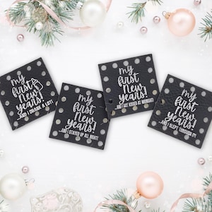 May include: Four black square coasters with white text that reads "my first New Years!" with different phrases below. The coasters are surrounded by Christmas ornaments and greenery.