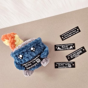 May include: A blue crocheted plush toy shaped like a campfire with a yellow and orange flame. The toy has a face with black eyes and a white mouth. The toy is surrounded by four black rectangular tags with white text. The text on the tags reads: "Emotional Support Dumpster Fire. It's fine. I'm fine. Everything's fine!" "Emotional Support Dumpster Fire. This fire really brings out the crazy in your eyes!" "Emotional Support Dumpster Fire. Burn it. Burn it ALL!" "Emotional Support Dumpster Fire. We didn't start the fire. It was ALWAYS burning."