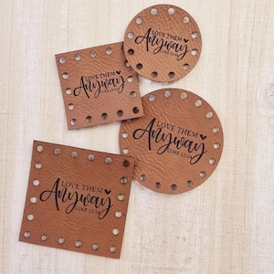 May include: Four brown leather patches with holes around the edges. Each patch has the text "LOVE THEM Anyway LUKE 23:34" in black lettering with a small heart symbol.