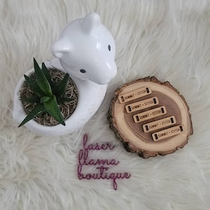May include: Five wooden tags with the text "Summit + Stitch" on a wooden slice. The tags are arranged in a stack, with the top tag being the most visible. The wooden slice is sitting on a white fluffy surface. There is a white ceramic llama planter with a green succulent plant in the background. The text "faser flama boutique" is written in pink glitter on the white surface.