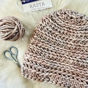May include: A chunky knit hat in a soft beige hue, accompanied by a matching ball of yarn and vintage-style metal scissors. A product label at the top displays the text RASTA, indicating super bulky yarn, with a weight of 690 grams.