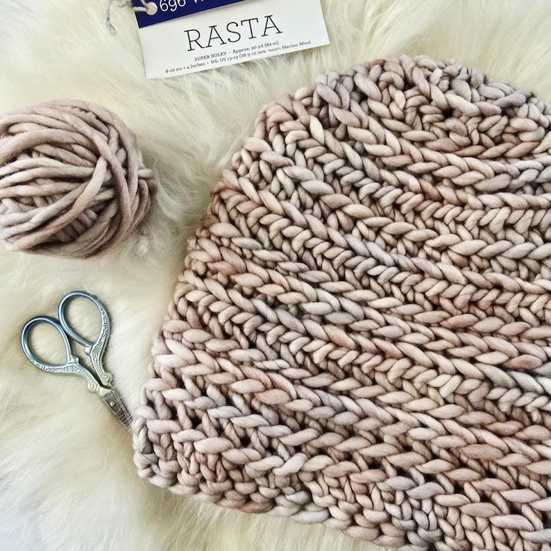 May include: A chunky knit hat in a soft beige hue, accompanied by a matching ball of yarn and vintage-style metal scissors. A product label at the top displays the text RASTA, indicating super bulky yarn, with a weight of 690 grams.