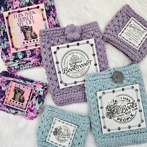 May include: A collection of handmade crochet book sleeves in various colors, including purple and blue. Each sleeve features a printed label with book-related quotes and illustrations, such as "This Book Ruined My Life."