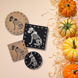 May include: Four coasters with a dog skeleton design, in various shapes: round, square, and a smaller square. The coasters are in black and tan colors. Decorative pumpkins and straw are in the background, suggesting a Halloween theme.