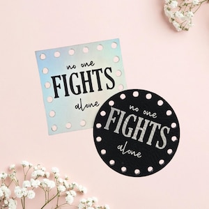 May include: A square iridescent patch and a circular black patch, both featuring pre-punched holes along the edges and the text "no one FIGHTS alone" printed in a combination of script and bold block lettering on the surface of each item.