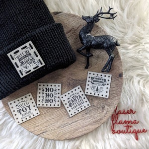 May include: A set of five wooden Christmas ornaments with black text. The ornaments feature phrases like "Meet me under the mistletoe", "Ho Ho Ho", "Santa's Toy Shop", "Santa Please Stop Here", and "Reindeer as they ride". The ornaments are on a wooden tray with a black reindeer figurine.