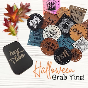 May include: A collection of round and square faux leather patches with pre-punched holes, featuring various Halloween-themed phrases and graphics, alongside a black rectangular tin labeled "hey boo" and several colorful autumn leaves.