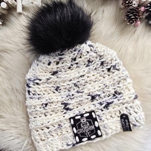May include: A chunky knit beanie in cream with dark gray flecks, topped with a large black faux fur pom-pom. A square black patch on the front features a white graphic of a nutcracker and the text CRUSHIN IT, with a small side tag reading LASER LLAMA.