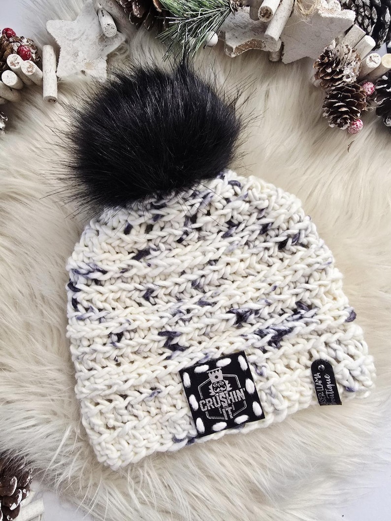 May include: A chunky knit beanie in cream with dark gray flecks, topped with a large black faux fur pom-pom. A square black patch on the front features a white graphic of a nutcracker and the text CRUSHIN IT, with a small side tag reading LASER LLAMA.