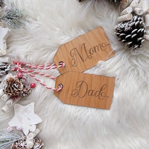 May include: Two wooden gift tags with the words "Mom" and "Dad" engraved in a cursive font. The tags are tied with red and white twine and rest on a white, fluffy surface. Decorative pinecones and greenery surround the tags.