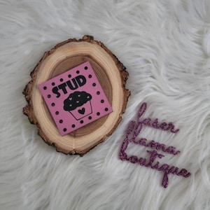 May include: A pink square with the word "STUD" and a cupcake illustration, surrounded by black dots, rests on a wooden slice. A glittery purple "laser llama boutique" logo is also present.