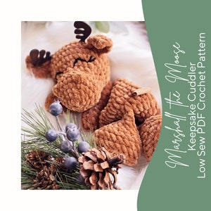 May include: A brown crocheted moose plush toy with closed eyes, lying on a white surface with a pine branch and pinecones. The text "Marshmallow the Moose Keepsake Cuddler Low Sew PDF Crochet Pattern" is in the background.