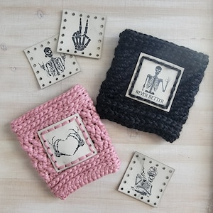 May include: Four square pieces of fabric with black skeleton designs and the text "Never Better". Two of the squares are attached to pink and black crocheted cup cozies.
