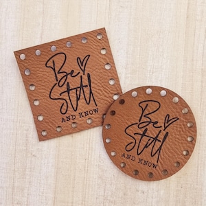 May include: Two brown leather patches with the phrase "Be Still and Know" in black lettering. The patches have holes around the edges.