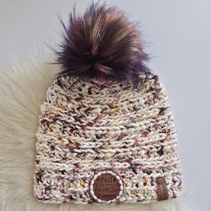 May include: A chunky knit beanie in a cream-colored yarn with flecks of purple, orange, and brown. It features a large, multi-colored faux fur pom-pom on top. A circular leather patch reads "Fall is my second favorite F-word" and a small tag says "Llama Boutique."