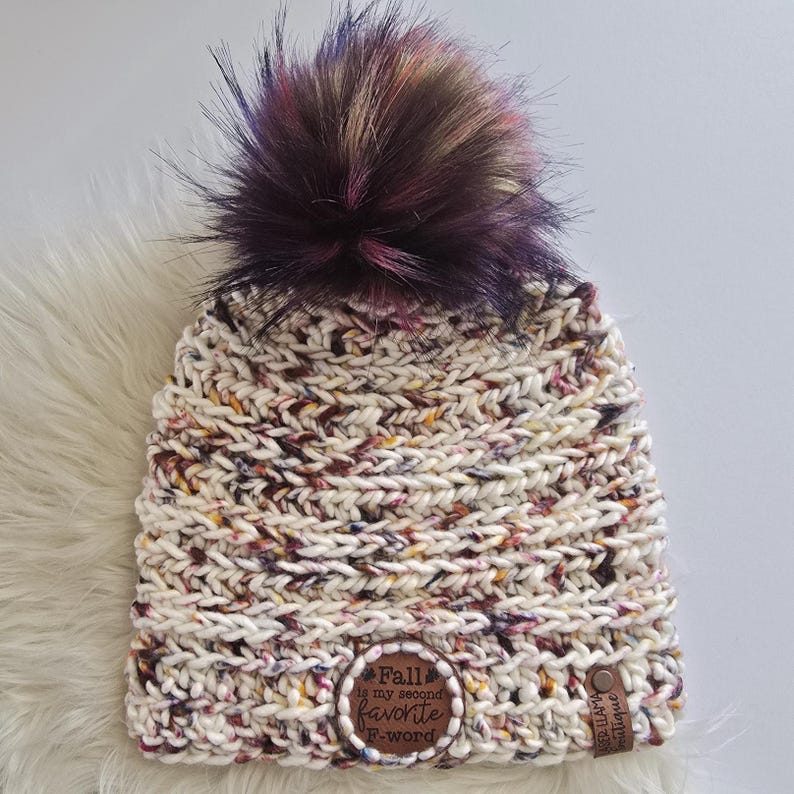 May include: A chunky knit beanie in a cream-colored yarn with flecks of purple, orange, and brown. It features a large, multi-colored faux fur pom-pom on top. A circular leather patch reads "Fall is my second favorite F-word" and a small tag says "Llama Boutique."