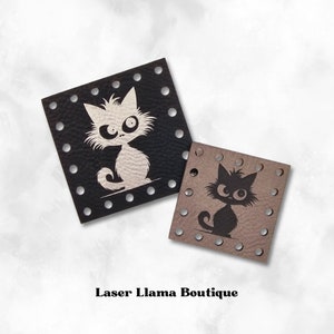 May include: Two leather patches featuring cat designs. The larger patch is black with a white cat with large eyes. The smaller patch is brown with a black cat with large eyes. Both patches have holes around the edges.