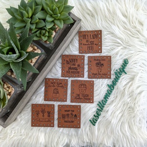 May include: Eight brown leather plant tags with various humorous sayings about plants. The tags have a dotted pattern around the edges and are displayed on a white fluffy surface.