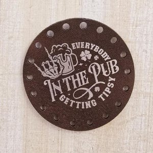 May include: Brown leather coaster with a white design that says "Everybody in the pub getting tipsy". The design includes a skeleton hand holding a beer mug and shamrocks.