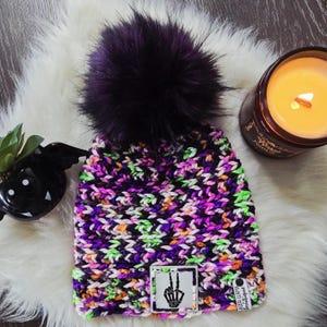 May include: A chunky knit beanie featuring a variegated yarn pattern in black, purple, orange, and neon green. The hat is topped with a large, dark purple faux fur pom-pom and includes a square patch displaying a skeleton hand peace sign.