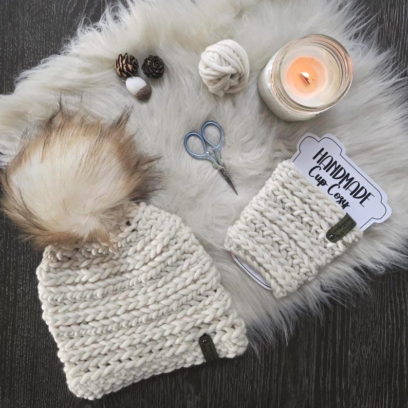 May include: A cream-colored chunky knit beanie with a large faux fur pom-pom and a matching cup cozy featuring a tag that reads Handmade Cup Cozy. The set includes a ball of yarn, small scissors, two pinecones, and a glass candle in a jar.