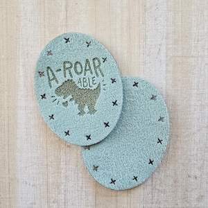 May include: Two oval, pale blue coasters. One has a dinosaur graphic and the text "A-ROAR-ABLE", with black cross decorations. The other is plain, with black crosses around the edge. Coasters are made of a soft material.