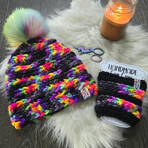 May include: A chunky knit beanie with a rainbow faux fur pom-pom and a matching cup cozy. Both items feature a pattern of black yarn alternating with vibrant neon pink, yellow, orange, and purple yarn. A small branded tag is attached to each piece.