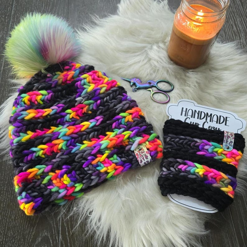 May include: A chunky knit beanie with a rainbow faux fur pom-pom and a matching cup cozy. Both items feature a pattern of black yarn alternating with vibrant neon pink, yellow, orange, and purple yarn. A small branded tag is attached to each piece.