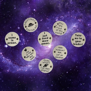 May include: Seven circular beige tags with two holes each, featuring black text and space-themed graphics. Phrases include Greetings Earthling, I come in peace, I just need more space, You are out of this world, Take me to your leader, Love you to the Milky Way and back, Alien invasion? Nah, just on vacation, and Guess what?! I've seen Uranus!