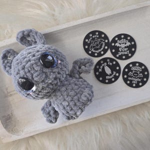 May include: A handmade gray crocheted bat plush toy with large, reflective blue eyes. The bat is displayed with four black circular tags with white text and bat illustrations. The tags read: "You are Fangtastic!", "Batty for You!", "Hang in There!", and "Just wingin' it!"