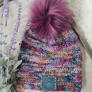 May include: A chunky knit beanie featuring a variegated yarn pattern in shades of purple, blue, and tan. The hat is topped with a large, fluffy dark magenta pom-pom and includes a square teal leather patch embossed with a ghost and floral design.
