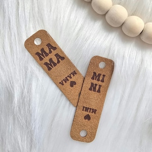 May include: Two brown leather tags with holes punched in the top. One tag says "Mama" in brown letters with a heart symbol and the word "Vinyl" below. The other tag says "Mini" in brown letters with a heart symbol and the word "Vinyl" below.