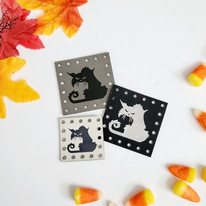 May include: Three black leather patches with a white cat silhouette design. The cat is sitting with its tail curled around its body. The patches are square and have holes around the edges.