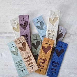 May include: A collection of rectangular fabric labels in various colors including blue, green, purple, and tan. Each label features an embroidered heart icon above the text "made with love" and a small horizontal line symbol at the top and bottom.