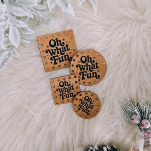 May include: Four wooden coasters with the phrase "Oh what Fun" in a retro font. Two are square, and two are round, all with small holes around the edges. The coasters are brown with black text and star accents.
