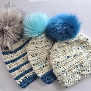 May include: Three chunky knit beanies with large faux fur pom-poms. The left beanie is cream with blue stripes, the middle is cream with a light blue pom-pom, and the right is cream with blue speckles and a dark blue pom-pom on top.