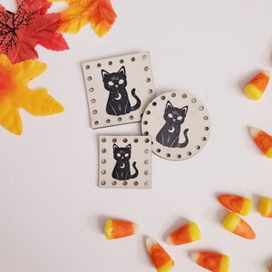 May include: Three wooden cat-shaped ornaments with a black cat design and a crescent moon. The ornaments are on a white background with candy corn scattered around them.