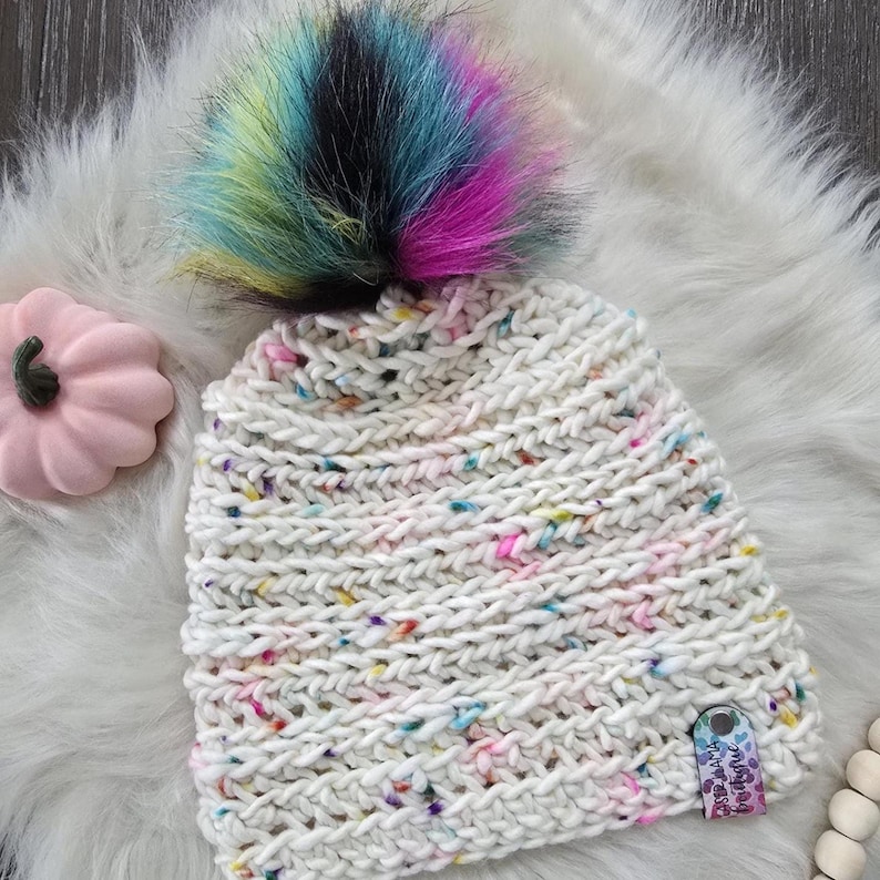 May include: A chunky knit beanie in off-white yarn with multicolored flecks, topped with a large, vibrant faux fur pom-pom featuring streaks of blue, yellow, pink, and black. A small rectangular tag with the text "Caster Mama Boutique" is attached.