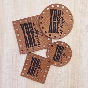 May include: Four brown leather patches with the text "YOU ARE enough" in black lettering. The patches are different sizes and shapes, with some having a heart symbol.