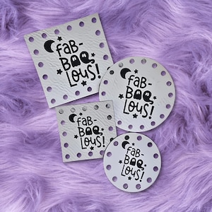 May include: Four white, textured coasters in different shapes, each featuring the black phrase "fab-BOO-lous!" with a crescent moon, stars, and a ghost. They are arranged on a fluffy, purple surface.